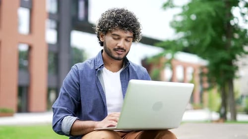 Young man using laptop while sitting on a bench on street near a modern building. Smiling handsome