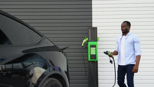 Black Man Plugging in Charging Cable to Electric Vehicle and Charges Batteries at Station