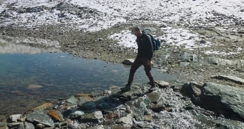 Male hiker crosses mountain stream by using stepstones, reflection
