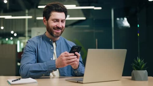 Male Office Worker Holding Smartphone and Looking at Screen at Desk with Laptop