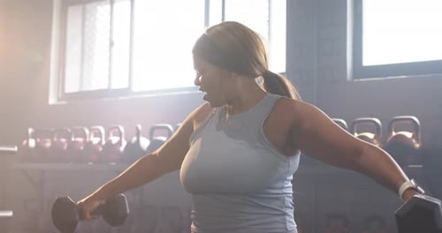 Gripping dumbbells, woman in gym raising laterally to shoulders, repeating for strength, smiling