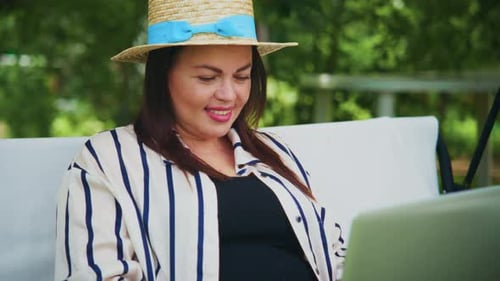 Woman in Striped Shirt and Straw Hat Working on Laptop Outdoors in a Serene Garden Setting