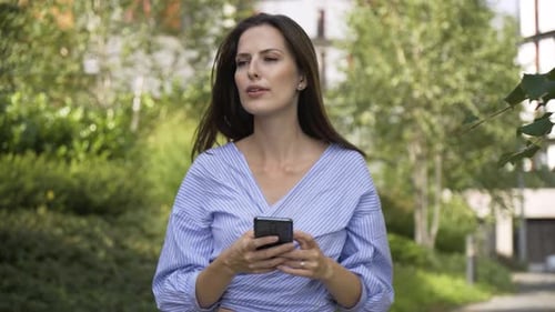 A Beautiful Young Caucasian Woman Works on a Smartphone with a Smile As She Walks Down a Street