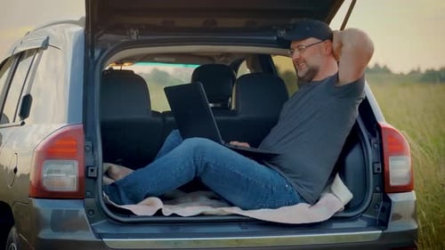 Homem feliz e sorridente trabalhando remotamente na natureza usando um laptop sentado no porta-malas do carro enquanto