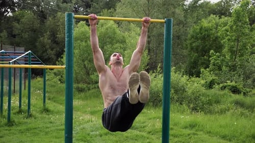 Athletic Man Trains Abdominal Muscles Raising Legs on Horizontal Bar at Sports Ground Young