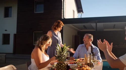 Group of adult friends having diner by swimming pool in sunny backyard.