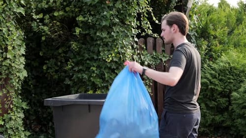 Caucasian male taking out trash in plastic bag to bin in garden