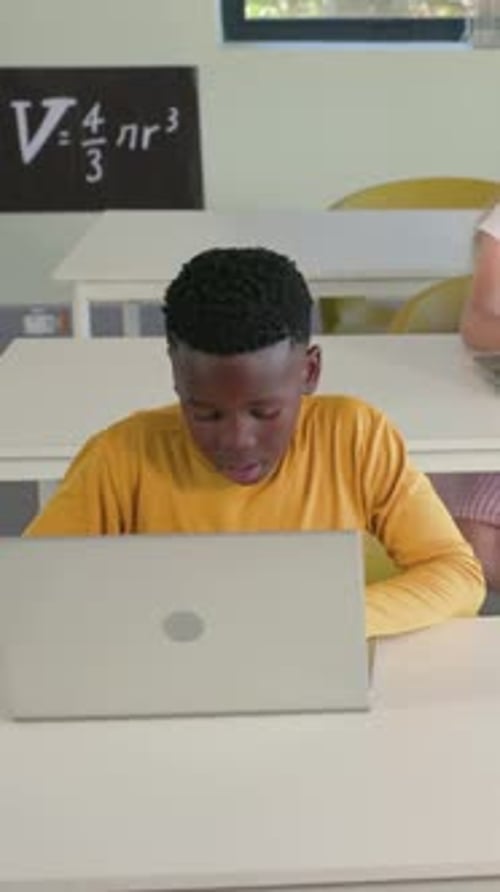 Vertical video: Working on laptop, student in mustard top raising right hand at desk to get help