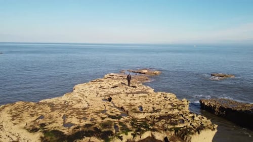 4K drone tracking shot of subject (person) walking towards camera, along rocky shoreline.