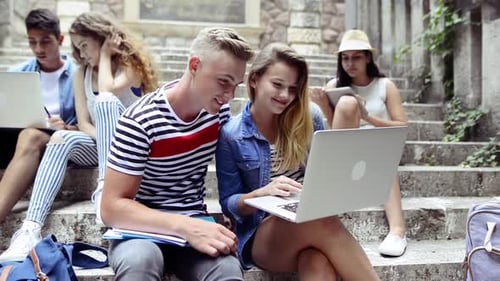 Trendy teenage students use gadgets on stone steps outside university building