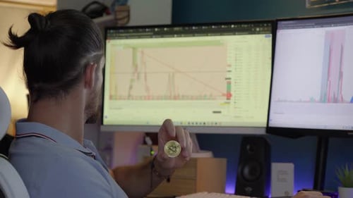 close-up shot of a man holding a Bitcoin whilst looking at crypto graphs