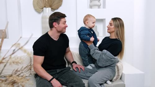 Mother plays with her cute little son learning to stand. Dad sits beside his loved ones smiling.