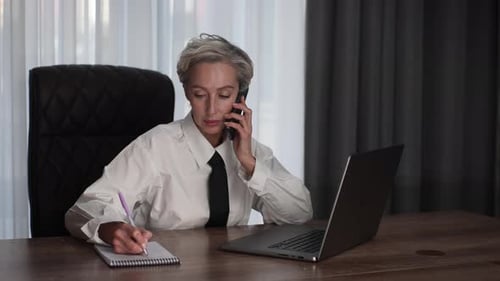 Busy Middleaged Business Woman Multitasking in Office Working Online with Laptop Talking Mobile