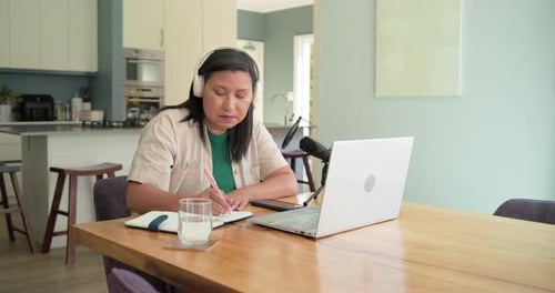Femme mature invitée par un appel, assise à la table de la maison, utilisant un ordinateur portable parlant dans le micro, notant
