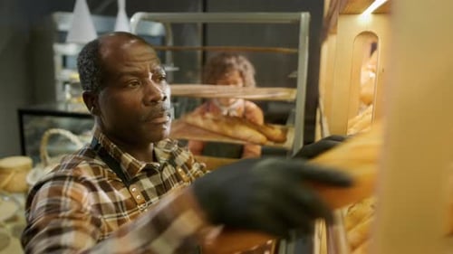 Black Bakery Worker Placing Fresh Baguettes on Bread Display for Sale