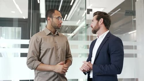Two businessmen shaking hands after agreement or deal while standing in a modern business office.