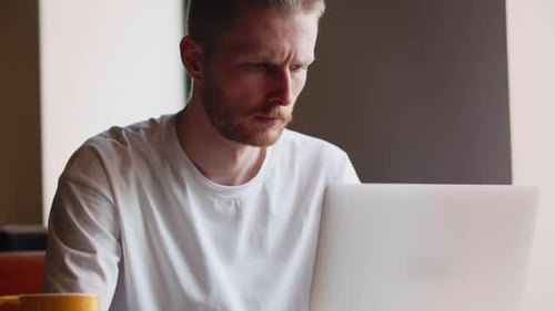 Freelancer Sits in a Cafe with a Cup of Coffee and Works on a Laptop A Frowning Man Looks at a