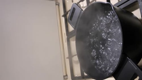 A Woman's Hand Opens the Lid of a Black Pot Where Water is Boiling