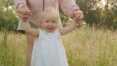 Smiling Toddler Infant Baby Walks Through Meadow in Green Grass in Park with Help Support of Mother