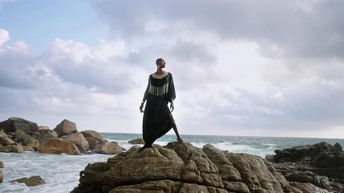 Godlike Gender Fluid Person of Color in Luxury Dress Jewelry Stands on Cliff Peak at Scenic Ocean