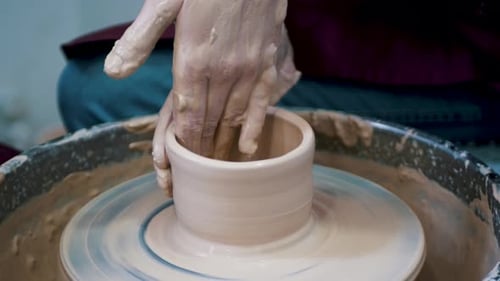 Close-up of potter's hands covered with clay making beautiful vase on throwing wheel in pottery work