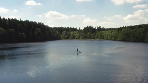 A lone person paddling a stand up paddleboard across a calm pond on vacation, aerial orbit
