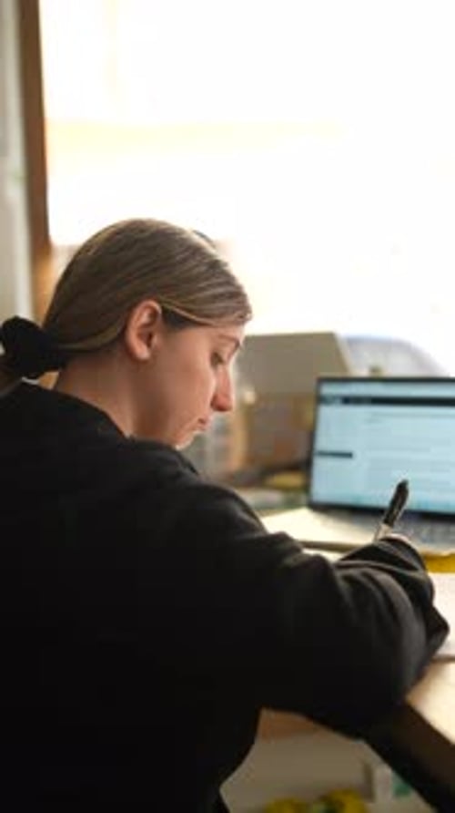A Woman is Working and Taking Notes at a Desk with a Laptop While Drinking Coffee