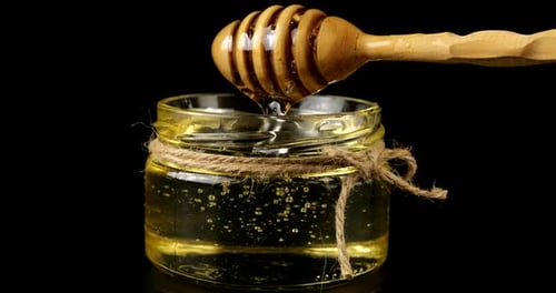 Honey flows down from a spoon spindle into a jar with honey on a black background.