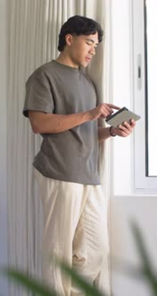 Homme en t-shirt se concentrant sur l'écran de la tablette à côté d'une fenêtre lumineuse, tapant et vérifiant les textes