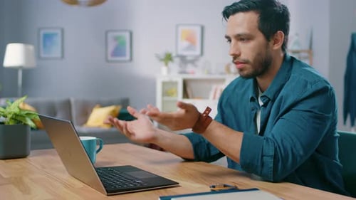 Handsome Young Man Makes Video Call with His Laptop Computer. His Sitting at the Desk in Cozy Livin