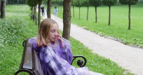 Teen Girl in Raincoat Sitting on Bench in Summer Park at Rainy Warm Weather