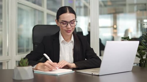 Business Lady Writing Information in Notebook and Working with Laptop in Office