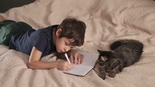 Boy Writes in Notepad with Kitten on Bed