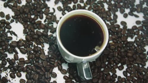 Rotierende Tasse Kaffee mit Dampf, umgeben von braun gerösteten Kaffeebohnen.