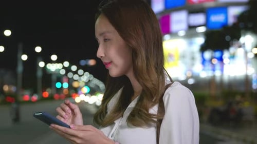 Молодая азиатская женщина улыбается и носит белую рубашку во время использования наушников со смартфоном на улице ночью