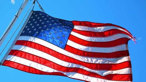 USA flag hanging on the mast flaps in the strong wind. Low angle view. Blue sky at backdrop.