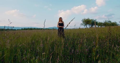 Beautiful female blonde model in long dress stands on a meadow in apple garden