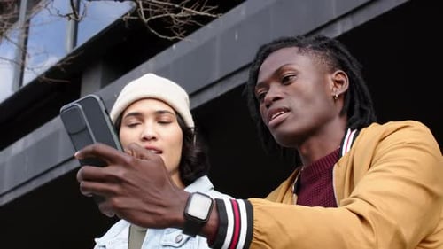 Diverse friends sitting on bench in plaza, glancing at smartphone then raising device for selfie