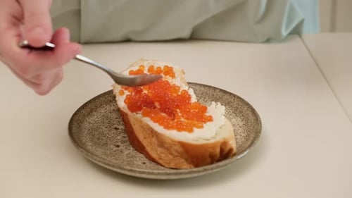 Slice of bread with butter and red caviar on ceramic plate. Hand holding spoon above sandwich. Close