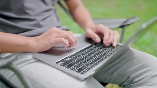 Freier Autor arbeitet in einem Park, Nahaufnahme Junger Mann sitzt auf einem Stuhl und arbeitet an einem Laptop