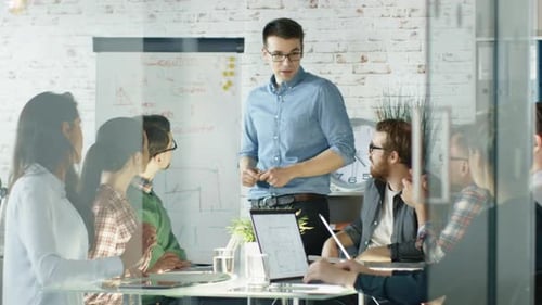 Man Makes Whiteboard Presentation to His Creative Office Staff. Coworkers Sit at the Big Glass Tabl