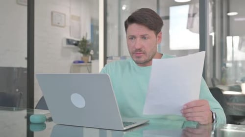 Man Reviews Paperwork and Laptop in Office Setting
