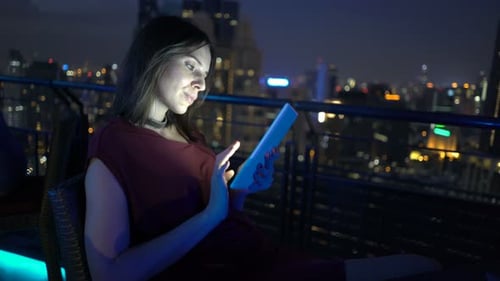 Young woman browsing the internet on tablet device at rooftop bar terrace at night