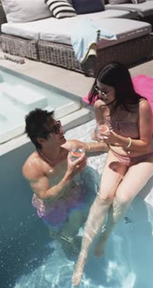 Vertical video: Raising man in trunks offering drinks to woman in pink swimwear at pool, toasting
