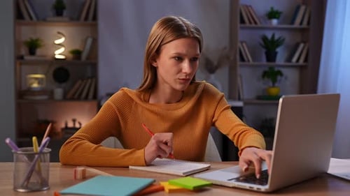 Woman Writing Notes at Desk with Laptop in Cozy Office Environment