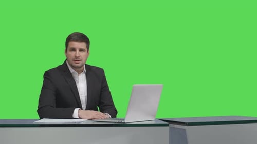 Media broadcaster is sitting at a table and talking on a mock-up green screen in the background.