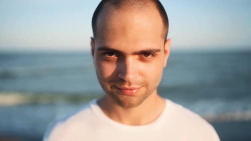 Portrait of Young Handsome Man Looking and Smiling at Camera Standing on the Beach with a Sea on