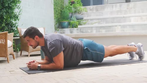 Mann legt auf der Terrasse in kurzen Hosen mit Matte Unterarme auf, hält ein Brett fest und spannt den Kern an, um Kraft zu gewinnen