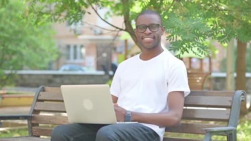 African American Man with Laptop Smiling at Camera while Sitting on Bench in Park