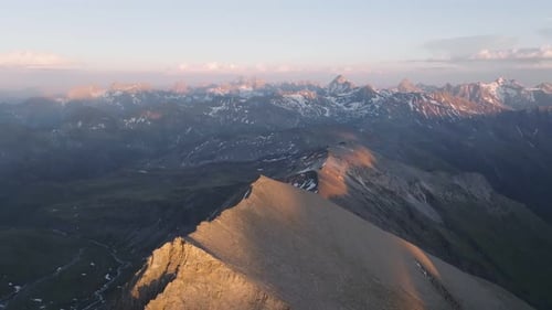Dramatic drone aerial of snow-flecked alpine ridge at sunset, descending into a deep mountain valley